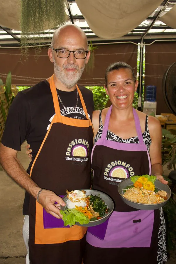 Two guests holding the plates they cooked, in daylight under the sail shades